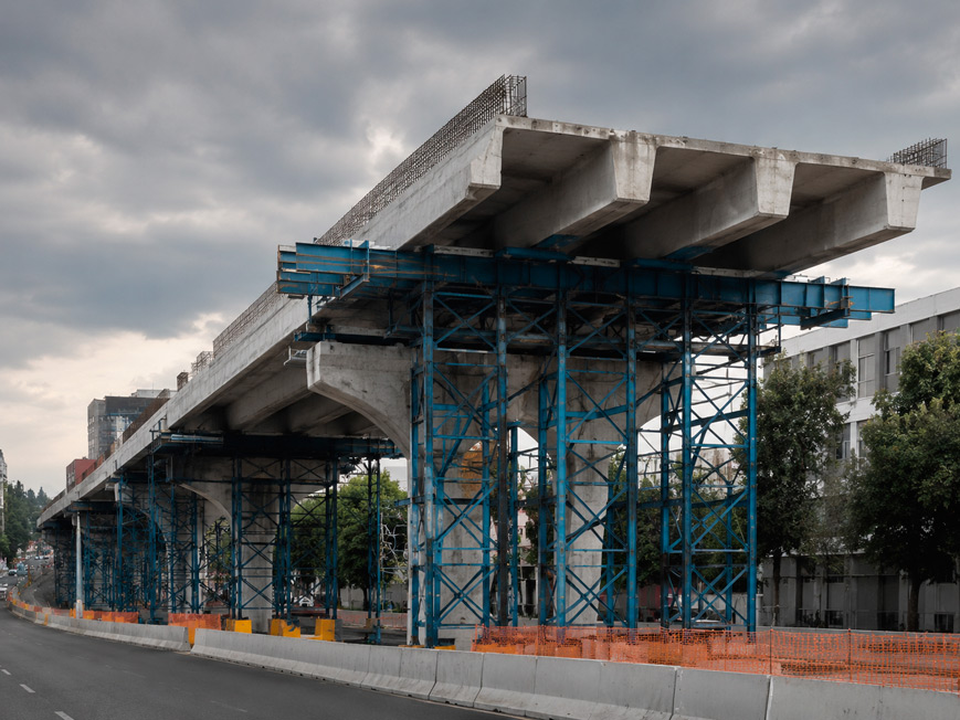 Fotografía documental de obra de ingeniería civil pesada en Ciudad de México: vista de frente de tramo de puente vehicular urbano en construcción con tablero de concreto hidráulico gris claro recién colado; trabes de concreto preesforzado grises visibles en la parte inferior del tablero; estructura de cimbra metálica azul oscuro soportando el tablero; calle de la ciudad al fondo con edificios y vegetación; cielo gris acero nublado; paleta: gris claro del concreto, azul oscuro de la cimbra metálica, negro asfalto, gris acero del cielo; composición de plano medio que muestra la estructura del puente en perspectiva desde el nivel de calle; sin personas visibles, sin texto, sin logotipos; estilo fotografía documental de gran infraestructura civil de alta producción.